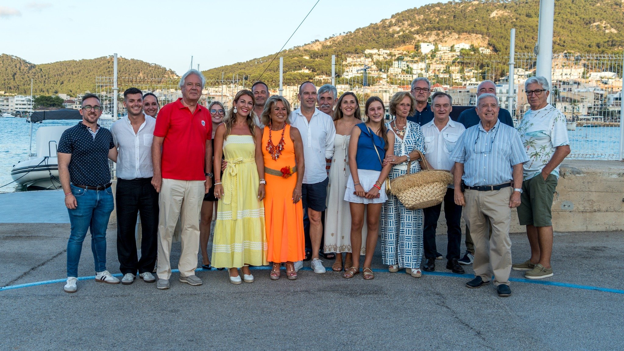 Grupo de personas posando en un muelle junto al mar, con colinas y edificios en el fondo.