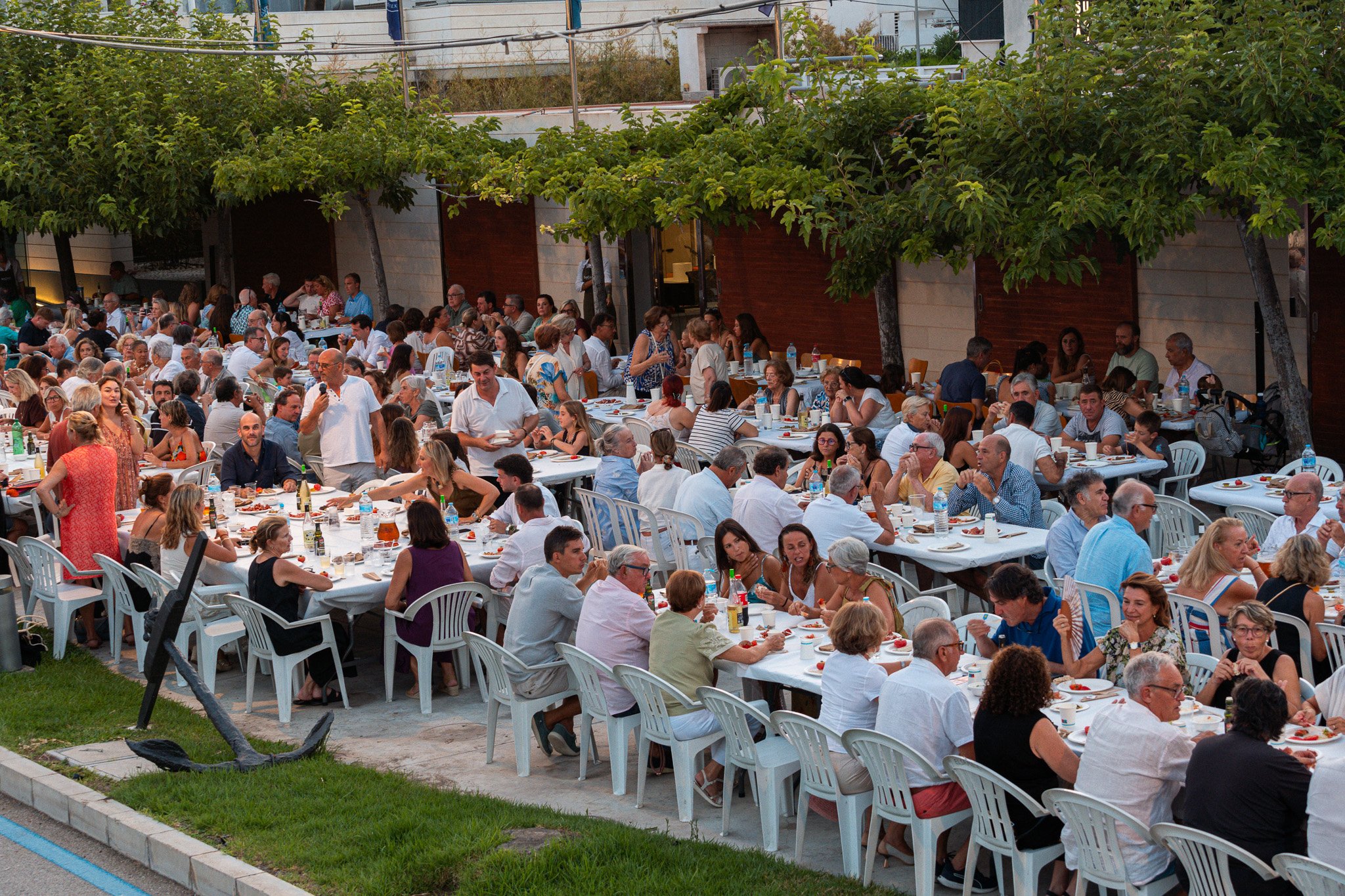 Una gran reunión al aire libre con muchas mesas y personas disfrutando de una comida. Hay árboles verdes y un fondo de edificios modernos.