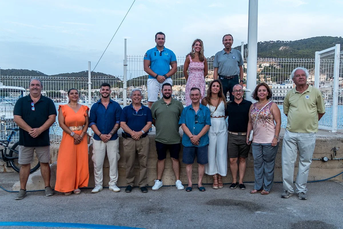 Grupo de personas posando frente a un parque con góndolas y montañas de fondo, algunos en la acera y otros en una estructura elevada.