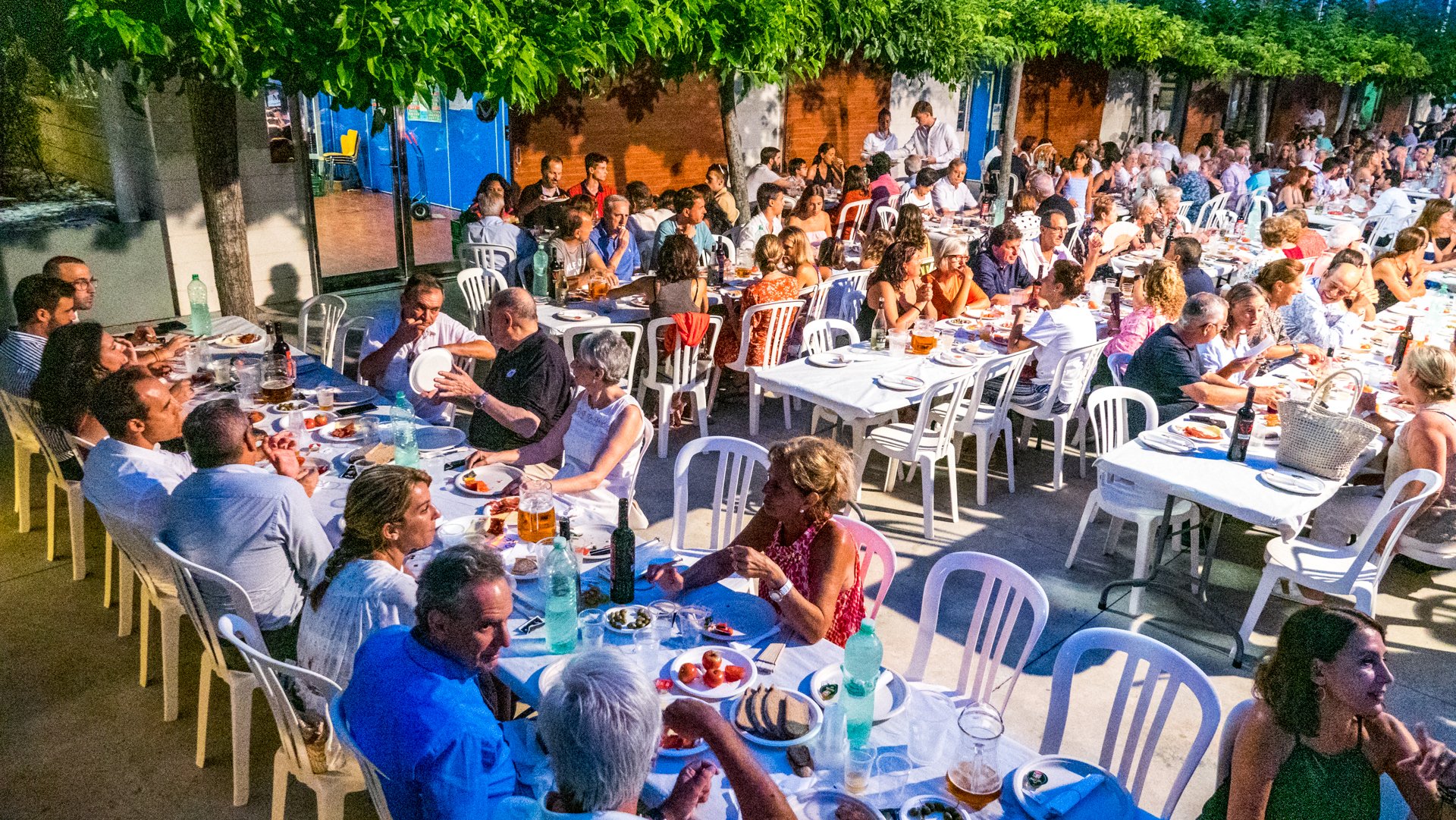 Una comida al aire libre con muchas mesas con personas sentadas comiendo y bebiendo, en un ambiente nocturno con árboles y paredes de ladrillo.