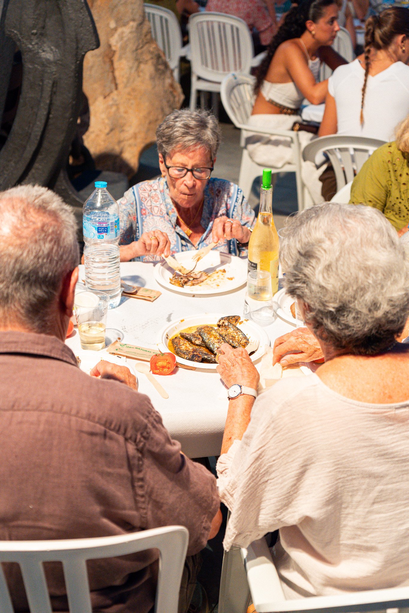 Varias personas adultas sentadas en una mesa en un restaurante, disfrutando de una comida, incluyendo sardinas y otros platos, con cinco en los primeros planos y varias en el fondo.