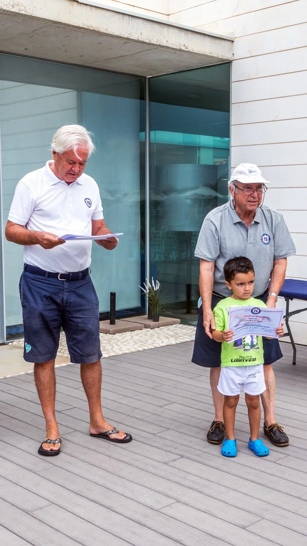 Tres personas, incluyendo un niño, en una ceremonia al aire libre. Dos adultos sostienen certificados, con un fondo de edificio moderno con paredes de piedra y vidrio.