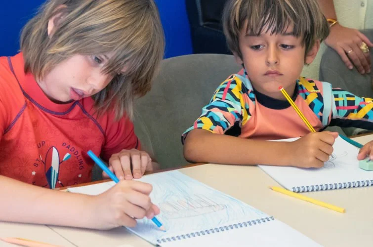 Dos niños niños pequeños sentados en una mesa, dibujando en cuadernos con lápices de colores.