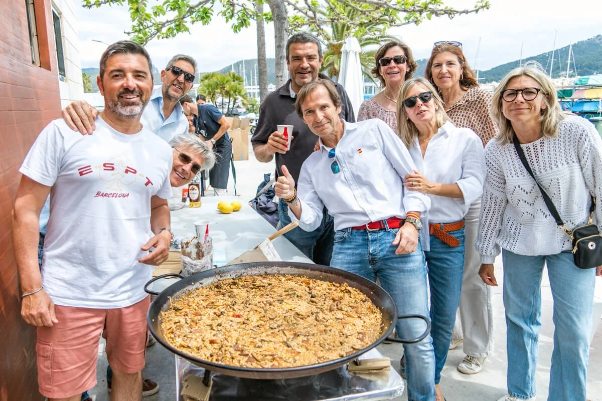 Grupo de personas reunidas alrededor de una gran paella en un evento al aire libre junto a un puerto, en un día soleado.