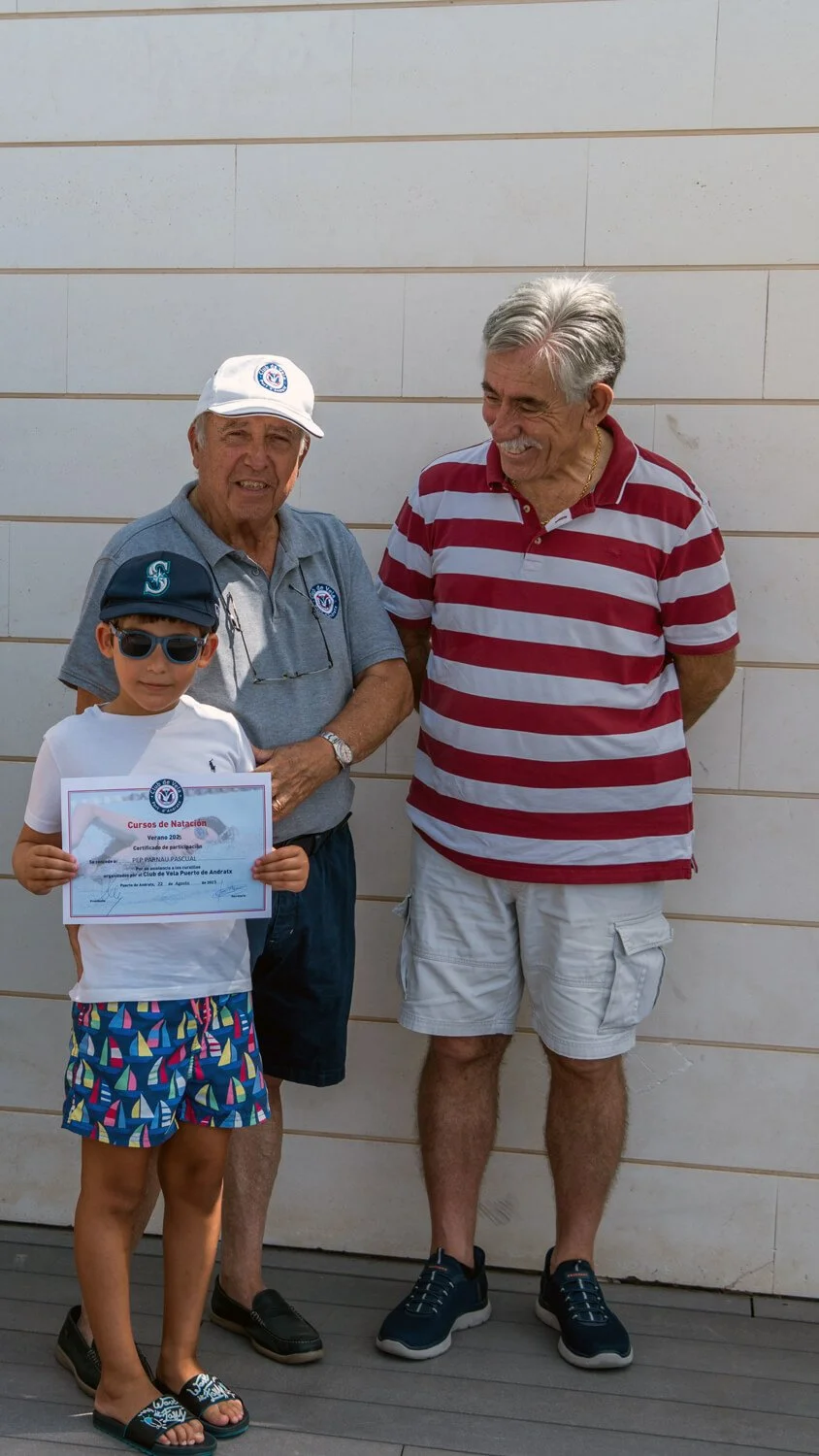 Un niño con gafas de sol y gorra sostiene un certificado, acompañado por dos hombres mayores sonriendo, uno con gorra blanca y otro con camiseta a rayas, en un fondo de pared blanca.