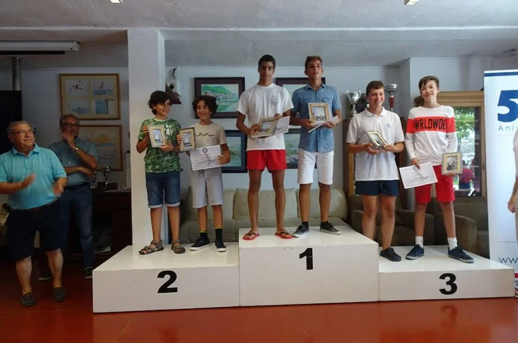 Niños en un podio de premios, con los primeros lugares en el centro, recibiendo trofeos y certificados, en una ceremonia de premiación, con adultos observando a su lado.