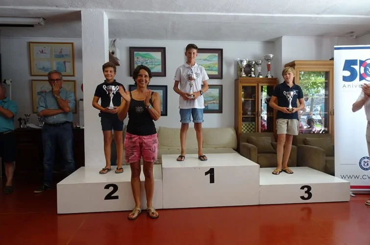 Niños en un podio de premiación con trofeos, en un evento de reconocimiento o competencia, con adultos y decoraciones en el fondo.