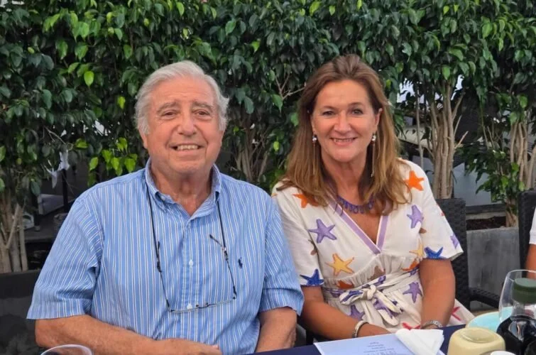 Hombre y mujer sentados en una mesa, sonriendo con fondo de arbustos verdes.
