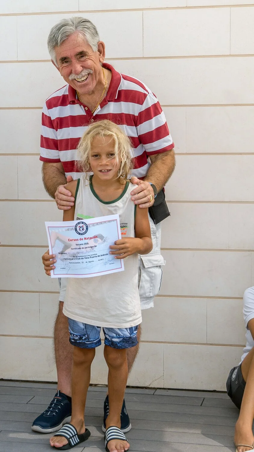 Un adulto y una niña en una cancha cubiert lapsúniendo a un niño con un diploma de participación en un curso de natación, el adulto viste una camiseta de rayas rojas y blancas y la niña usó camiseta sin mangas blanca y pantalones cortos con estampado