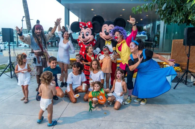 Niños y adultos en una fiesta con personajes de Disney, incluyendo Mickey Mouse y Minnie Mouse, en un espacio al aire libre.