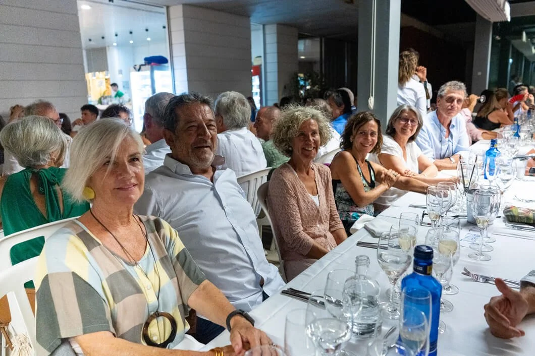 Grupo de personas sentadas en una mesa al aire libre, en una comida o cena social, disfrutando y sonriendo.