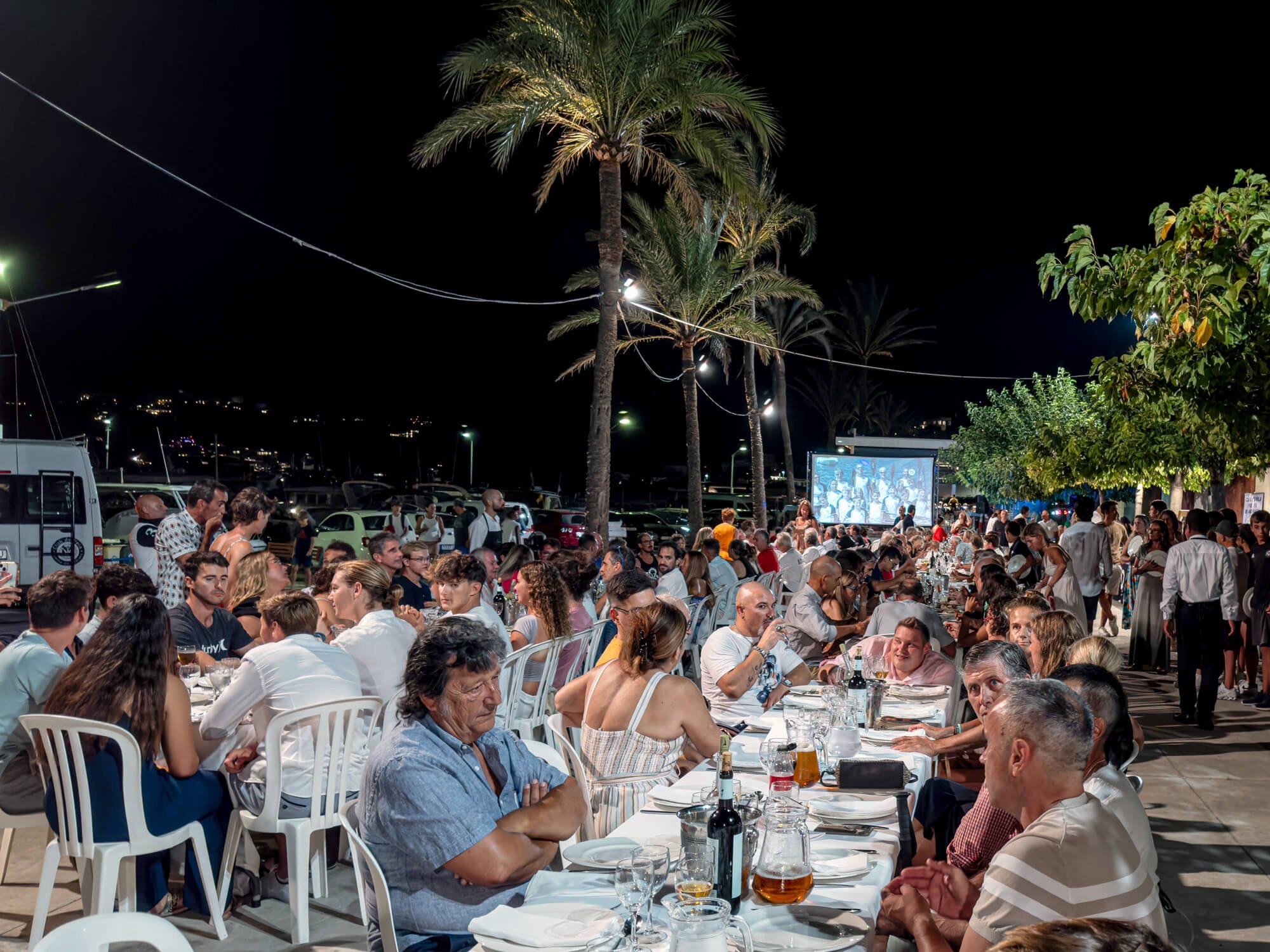 Una multitud de personas disfrutando de una cena al aire libre en la noche, con mesas con platos y botellas, árboles palmeras y pantallas de televisión.
