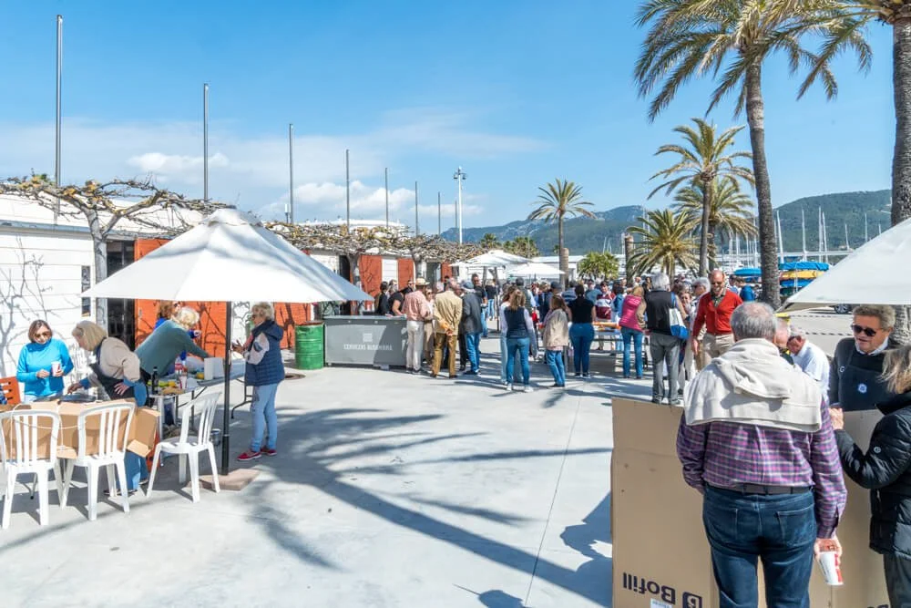 Imagen de una concurrida área al aire libre cerca de un puerto con palmeras, mesas con sombrillas, y muchas personas caminando y socializando.