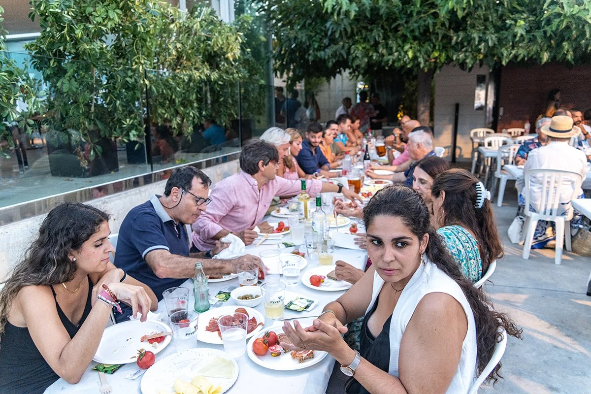 Grupo de personas cenando al aire libre en un restaurante, con árboles y plantas en el fondo.