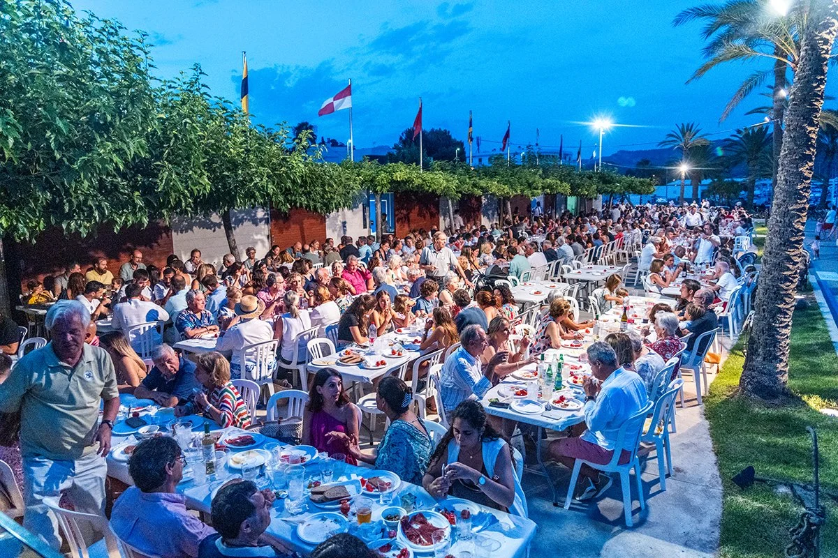 Gran reunión al aire libre con muchas mesas y sillas, gente comiendo y socializando, en un ambiente nocturno con palmeras y banderas en el fondo.
