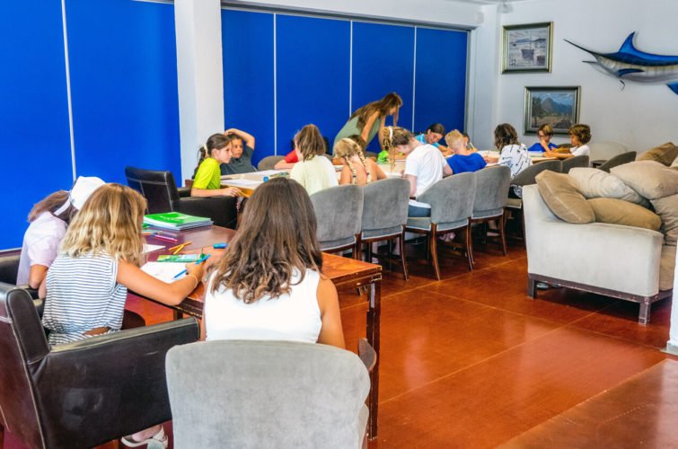 Grupo de niños en una sala, sentados en mesas, haciendo tareas o actividades, con una pared azul de fondo y decoraciones marinas en la pared.