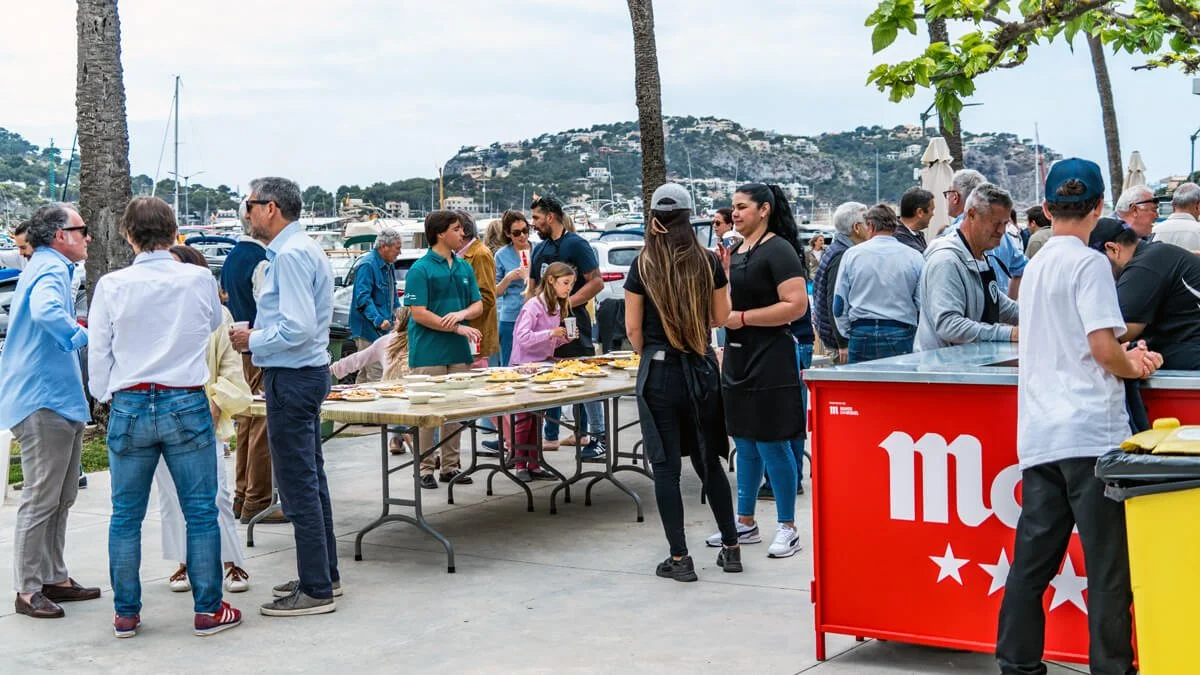 Personas en un evento al aire libre en un muelle, con mesas para comida, muchas personas conversando y un fondo de barcos y colinas.