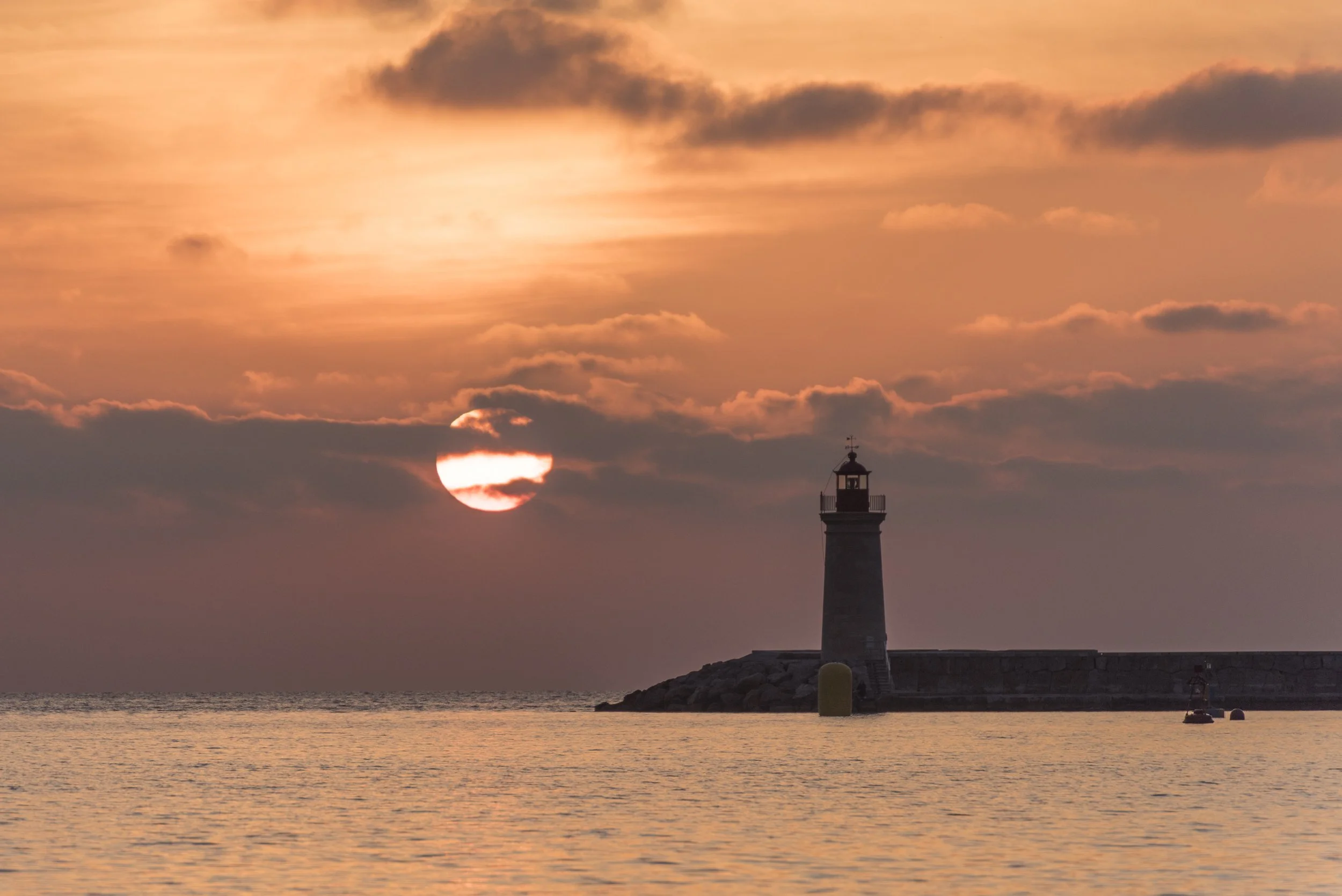 Atardecer sobre el mar con un faro en la orilla y el sol partially oculto tras las nubes en el cielo