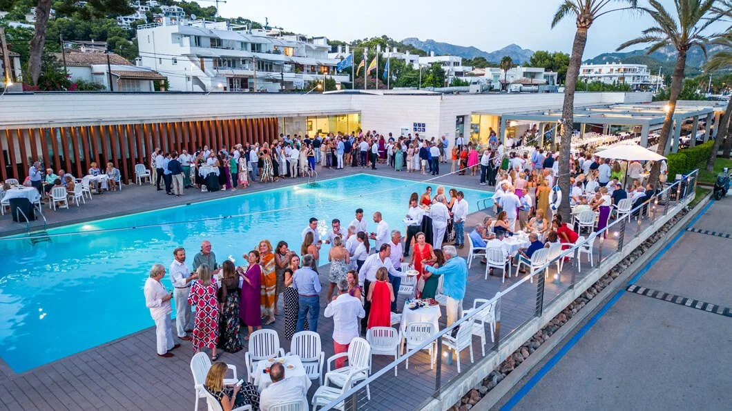 Celebración o evento social junto a una piscina ubicada en un área exterior con muchas personas reunidas, mesas con comida y bebidas, y un ambiente festivo en una zona con vegetación y edificios modernos al fondo.