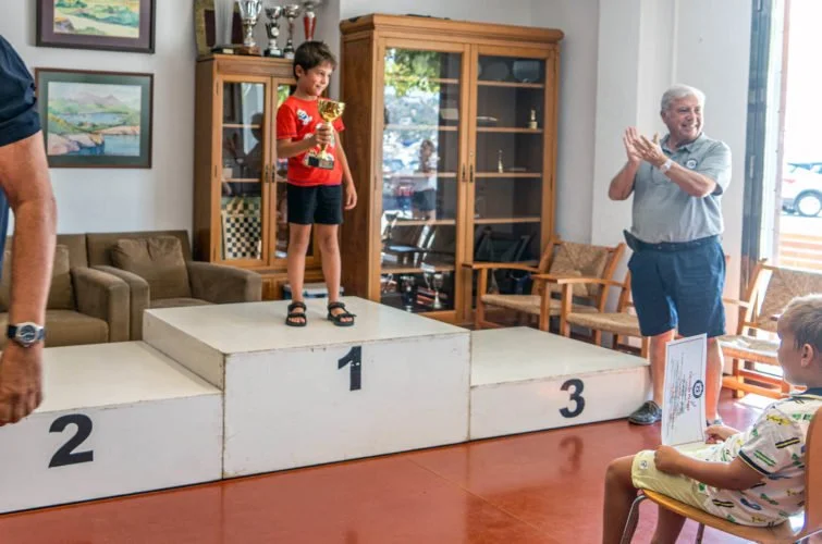 Niño en el primer lugar de un podio de premios, sosteniendo un trofeo, en una ceremonia de premiación en un lugar cerrado con un adulto y otros niños