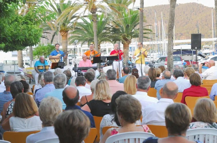 Un grupo de músicos tocando guitarra y otros instrumentos en un concierto al aire libre con audiencia adulta y palmeras de fondo.