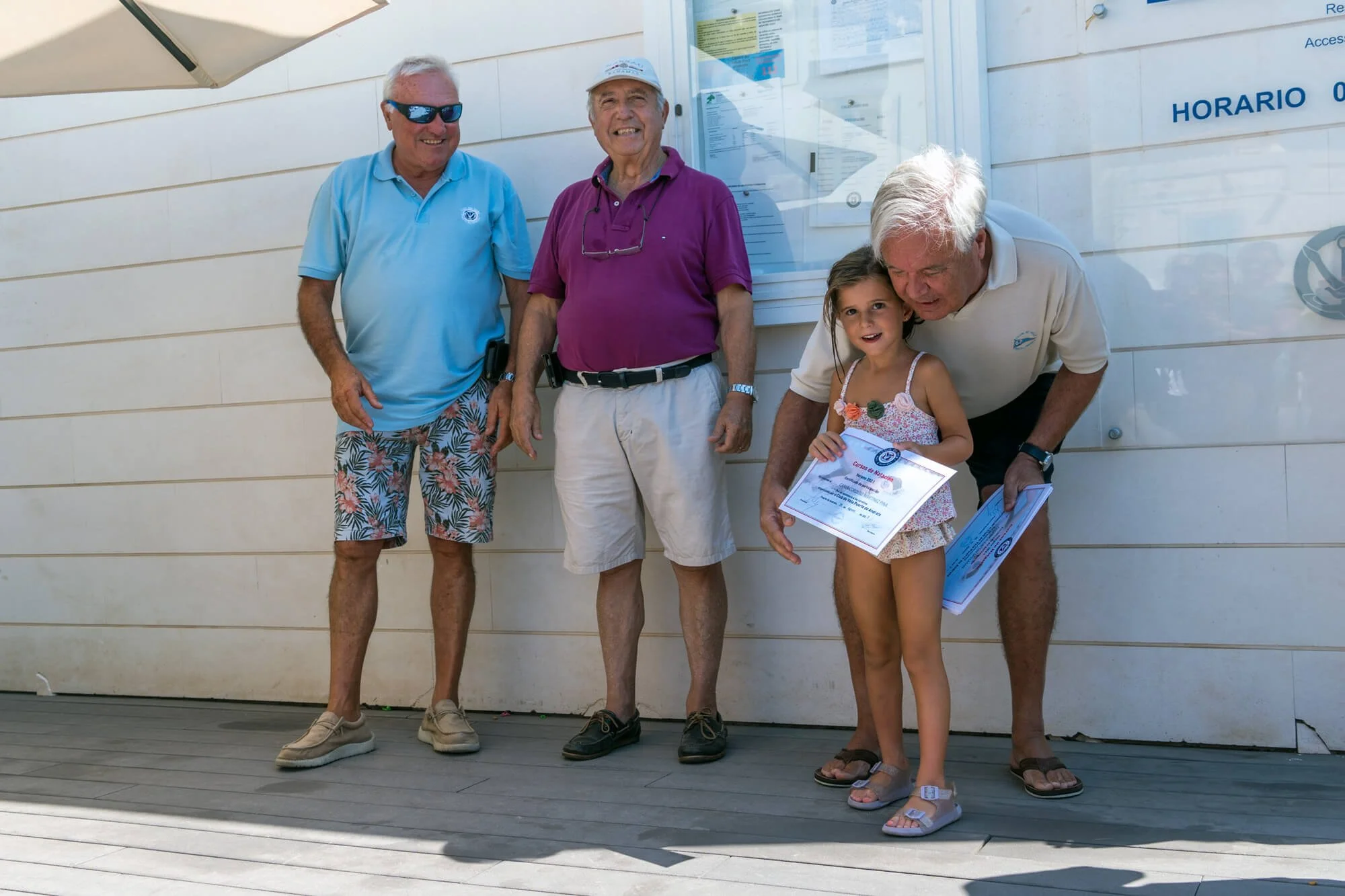 Grupo de adultos mayores y una niña sosteniendo certificados y papeles, de pie en un área exterior frente a un edificio con un cartel de horarios, disfrutando del sol en un día cálido.