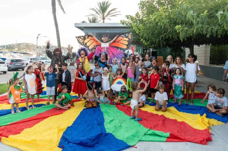 Niños y adultos disfrazados en una fiesta con decoración colorida y una mariposa gigante de fondo, en un espacio al aire libre con palmeras y autos estacionados.