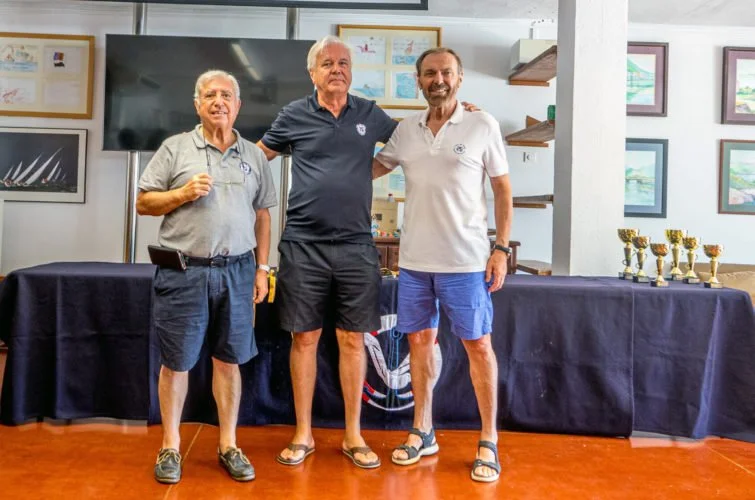 Tres hombres sonriendo y posando juntos en un evento con premios, en un entorno con cuadros colgados y una mesa con trofeos a su derecha.