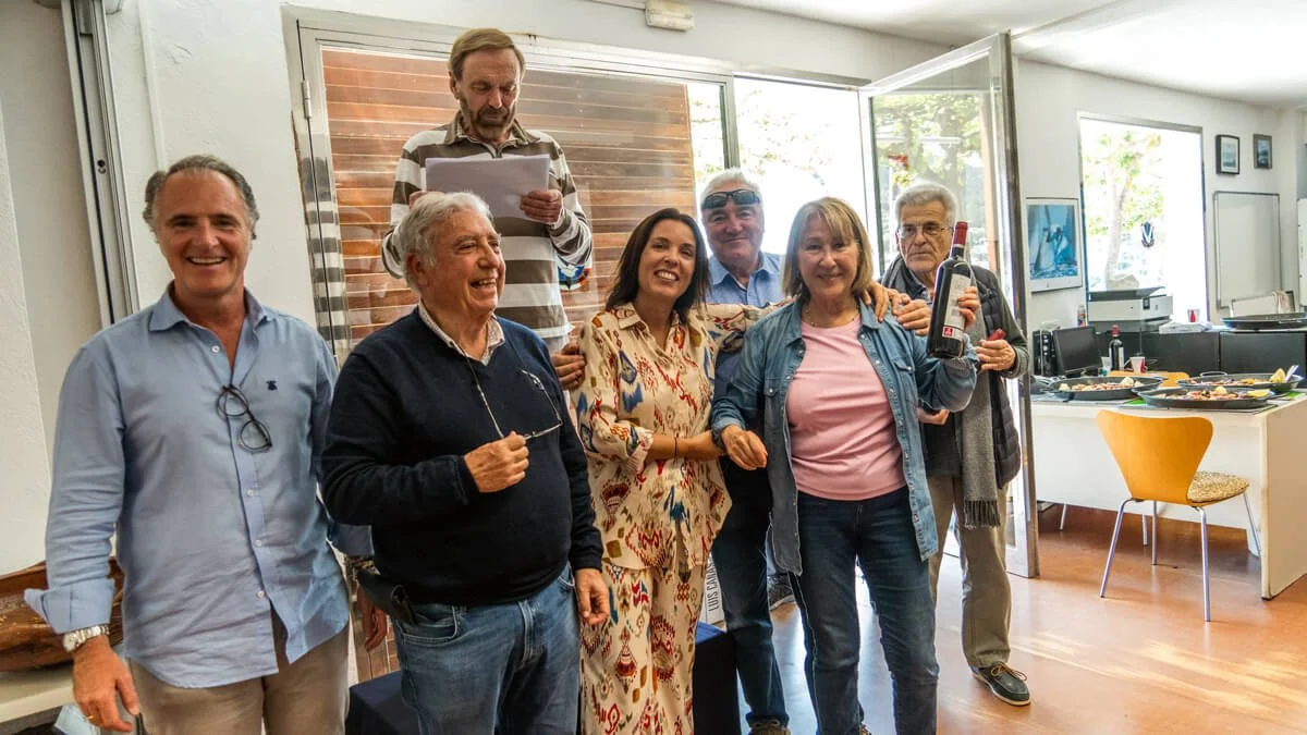 Grupo de personas en celebración, algunos con bebidas y sonriendo, en un ambiente interior con una mesa con comida en el fondo.