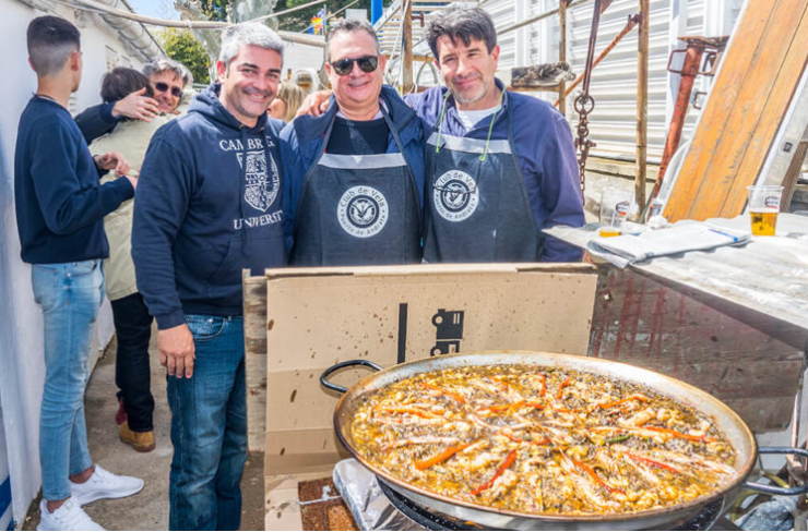 Tres hombres posando con una pizza en una feria o evento al aire libre.