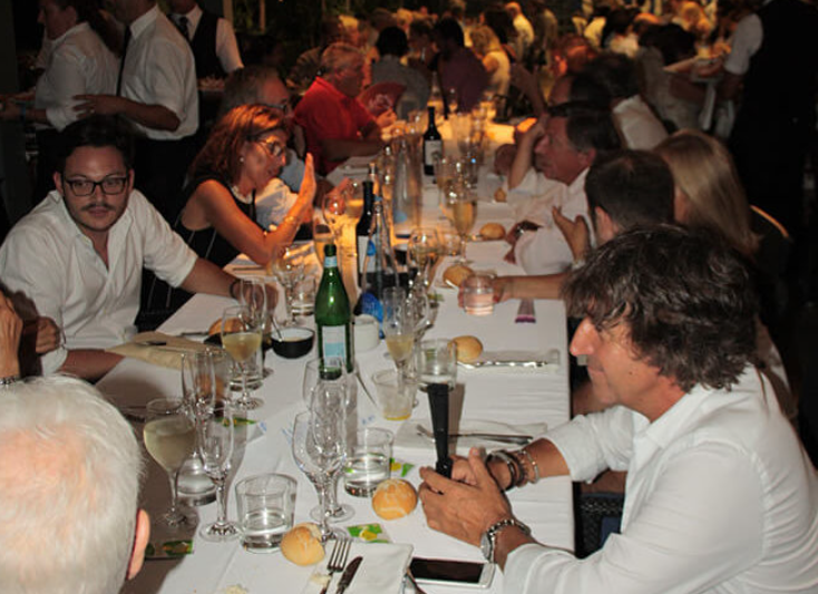 Mesa larga en un restaurante con muchas personas cenando, con copas, botellas y comida sobre la mesa, ambiente concurrido y elegante.