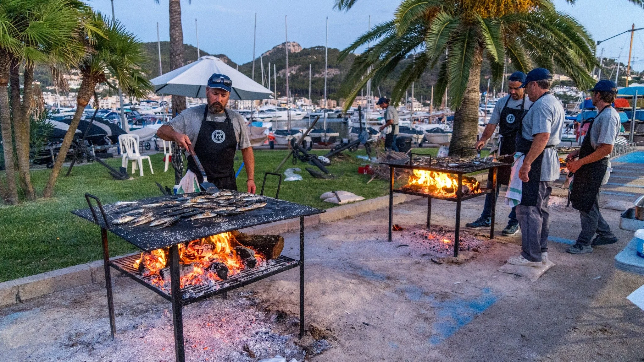 Personas cocinando pescado a la parrilla en un área al aire libre cerca de un puerto con barcos y montañas al fondo, en una tarde o noche.