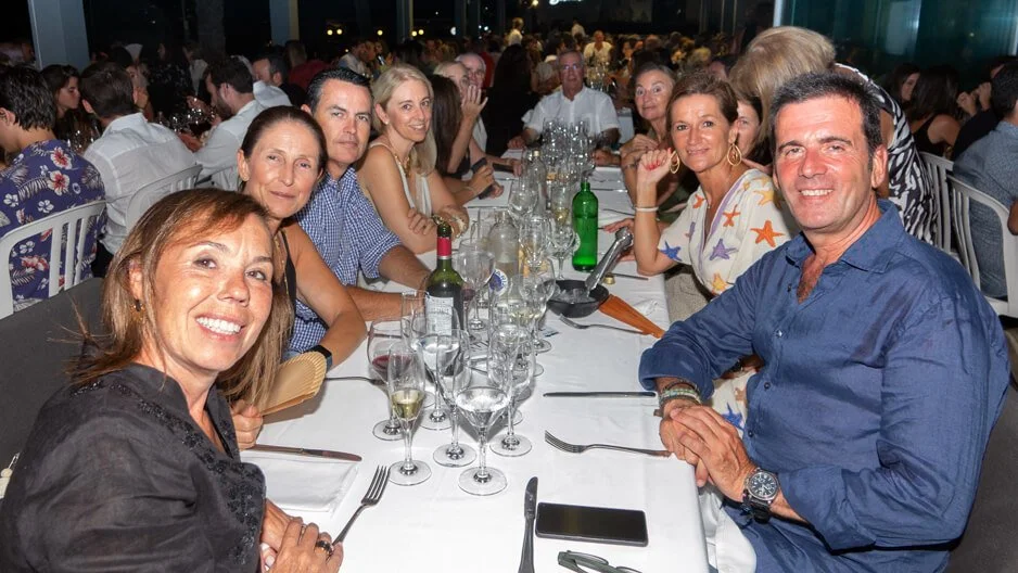 Grupo de personas sentadas en una mesa en un evento social, con comida, bebidas y varios invitados en el fondo.