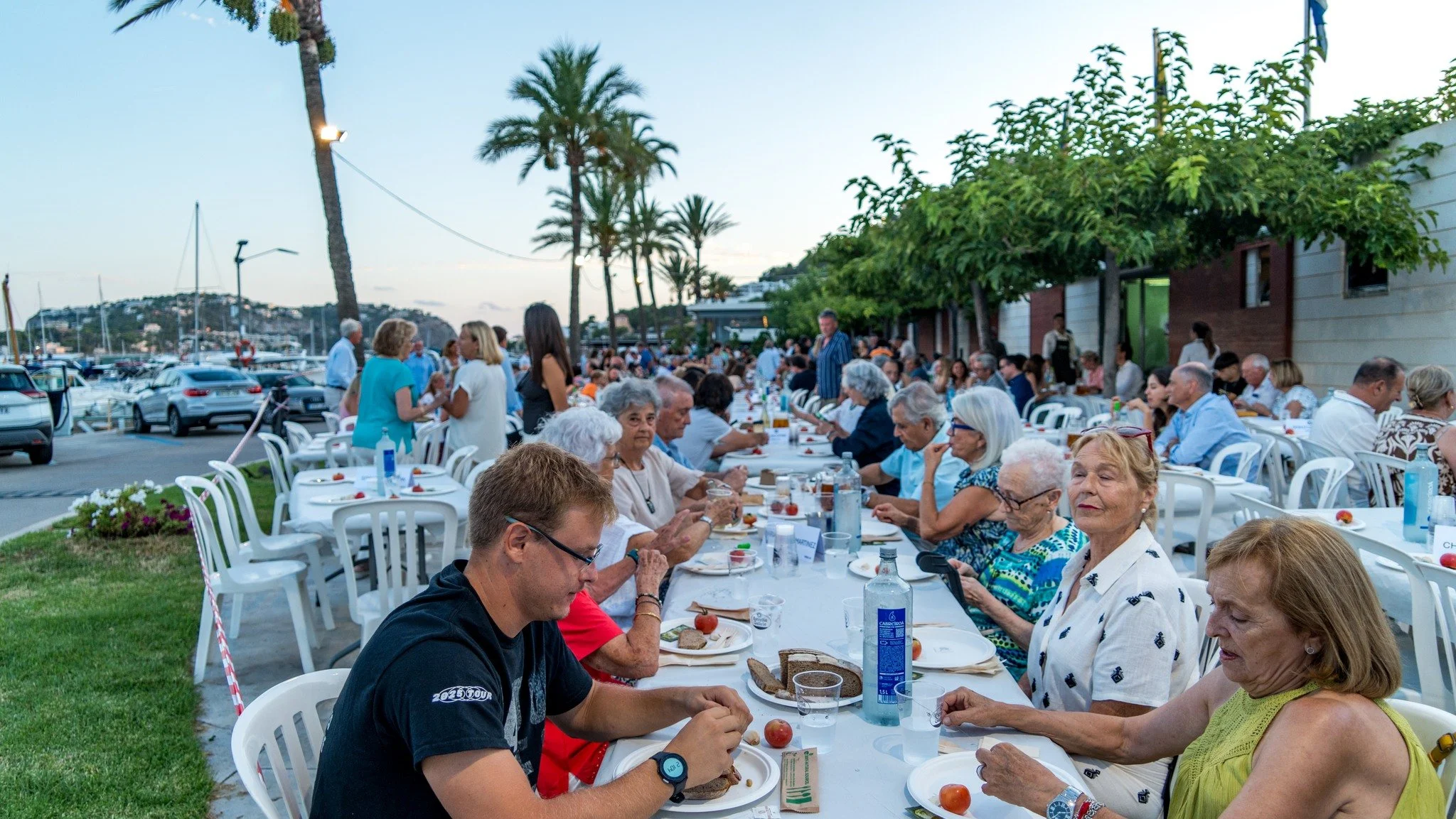 Una reunión al aire libre con muchas personas sentadas en mesas para comer, cerca de una marina con yates y palmeras, en la puesta del sol.
