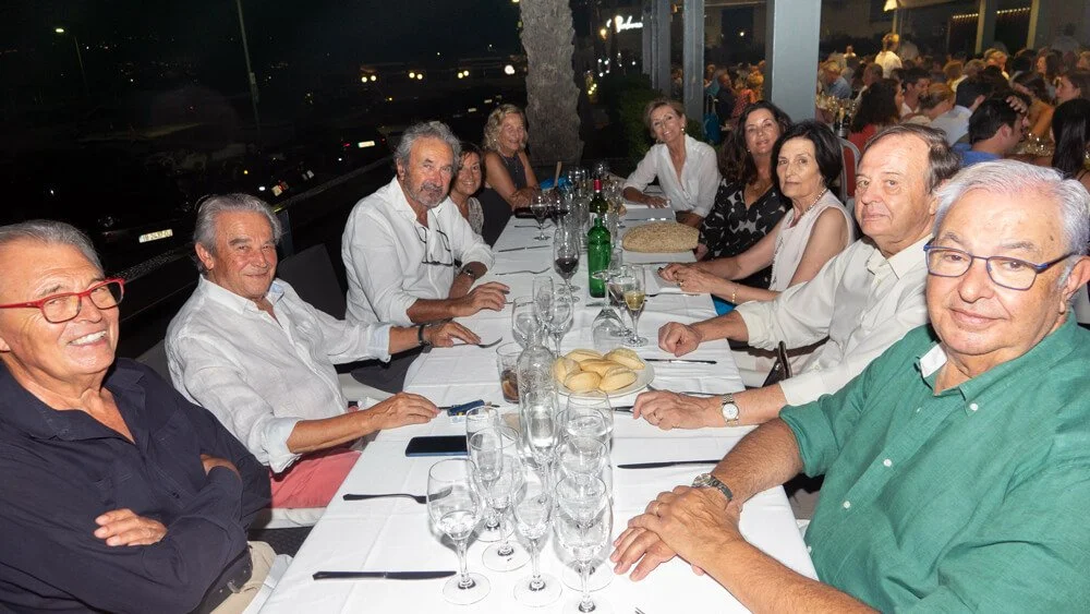 Grupo de personas sentadas en una mesa larga en un restaurante durante la noche, con varias bebidas y comida en la mesa.