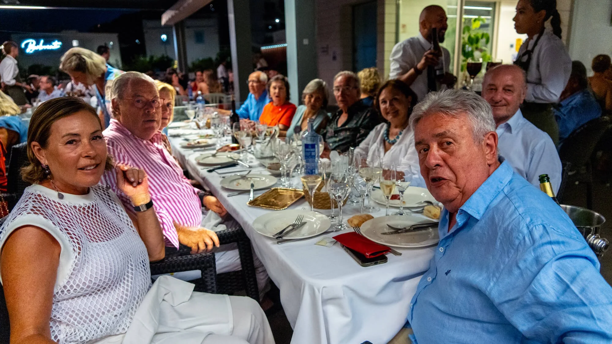 Grupo de personas sentadas en una mesa durante una cena al aire libre en la noche, con comida y bebidas en la mesa, en un ambiente festivo y social.
