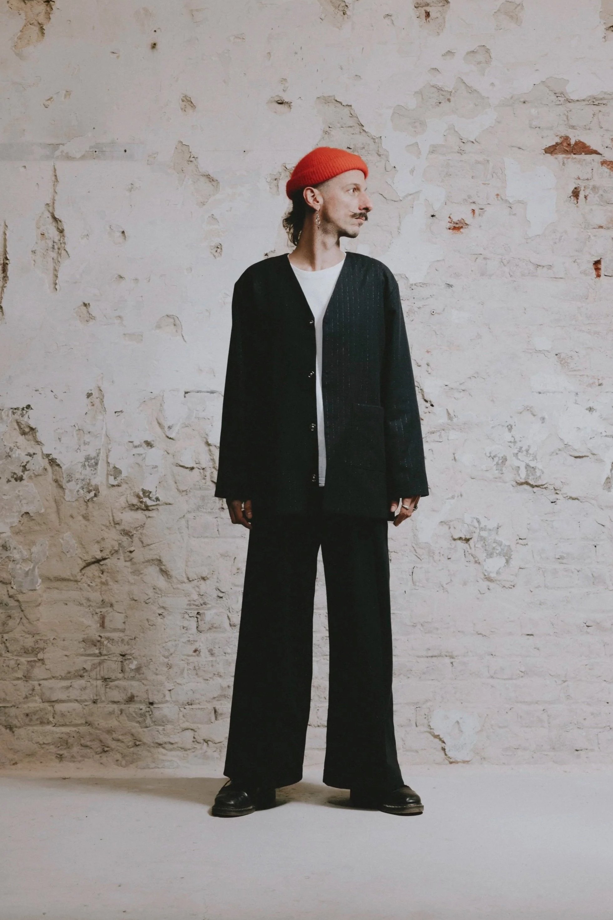 Un homme portant un béret rouge, un manteau noir et un pantalon large, debout devant un mur en pierre blanche.