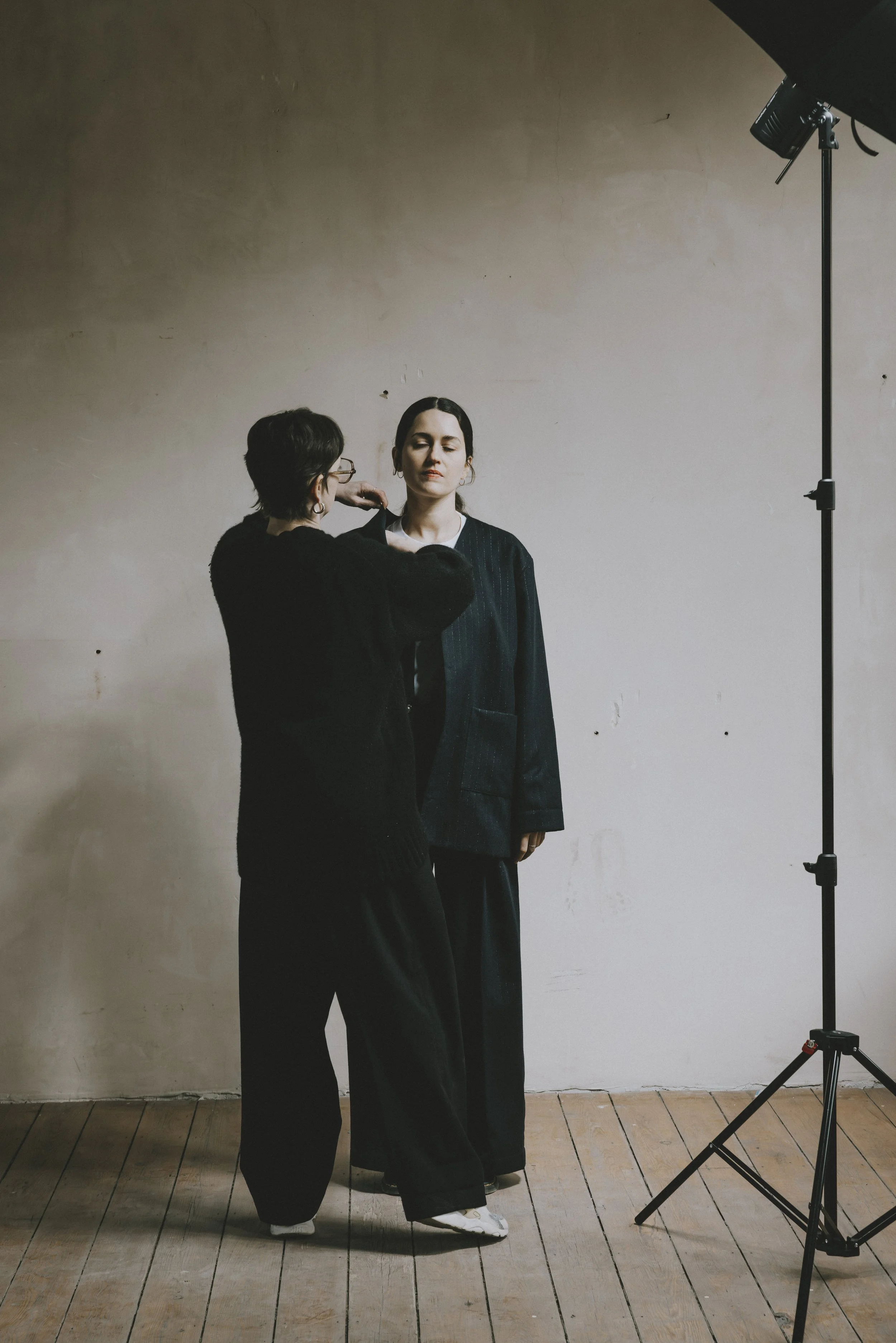 Une séance de photographie en intérieur avec deux femmes. L'une ajuste l'autre, qui porte une veste noire à rayures, contre un mur beige. Il y a un panneau de lumière sur un support à droite.