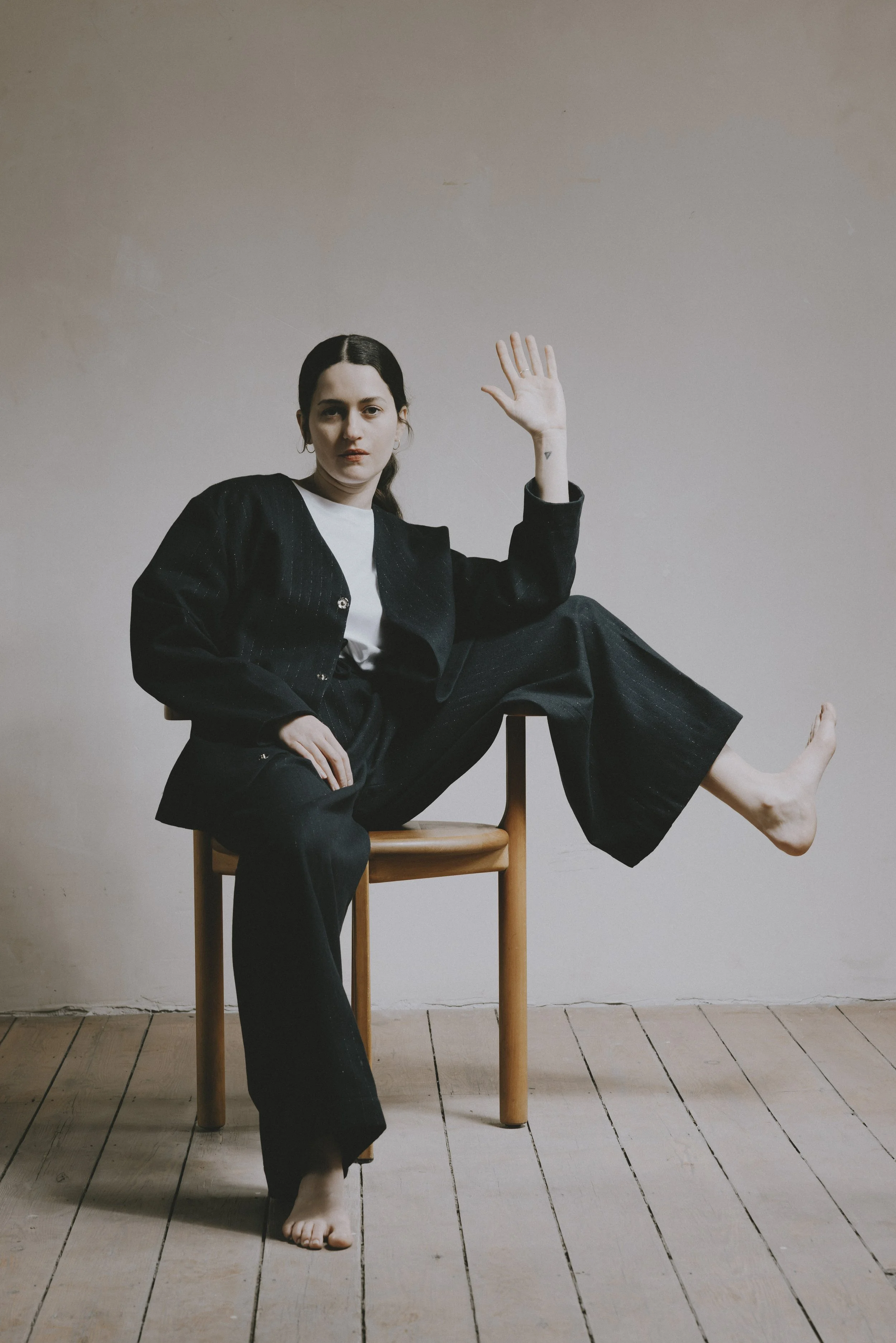 Une femme assise sur une chaise en bois, avec une jambe étendue sur son siège, portant une veste noire et un pantalon noir, le tout contre un mur clair, le sol en bois, la femme fait un signe de main levée.