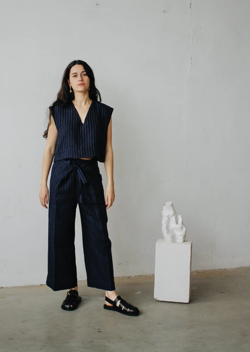 Femme debout portant un pantalon et un haut rayés noirs, chaussures noires, à côté d'une sculpture blanche sur un socle, dans un espace intérieur minimaliste.