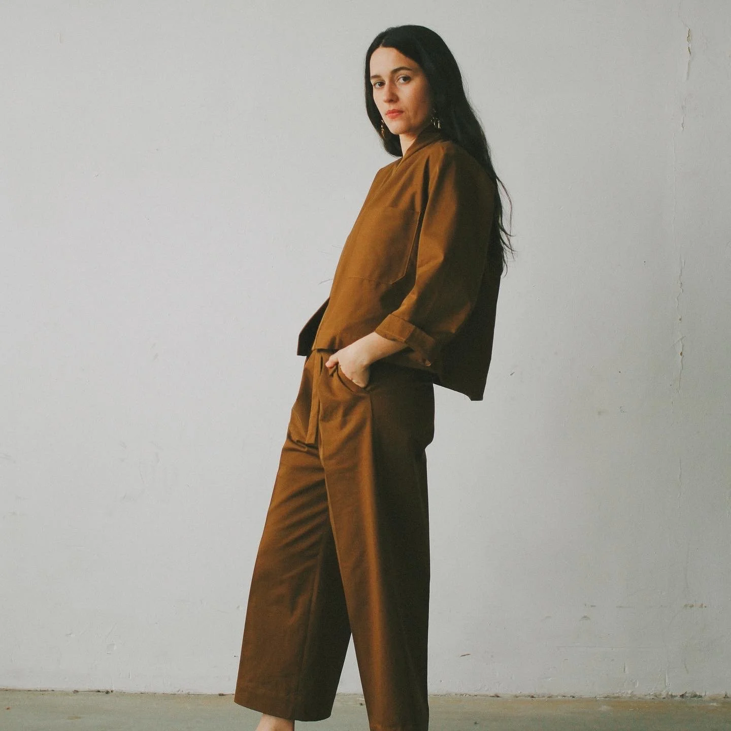 Femme portant un ensemble marron, vêtue d'une veste et d'un pantalon, se tenant contre un mur blanc.