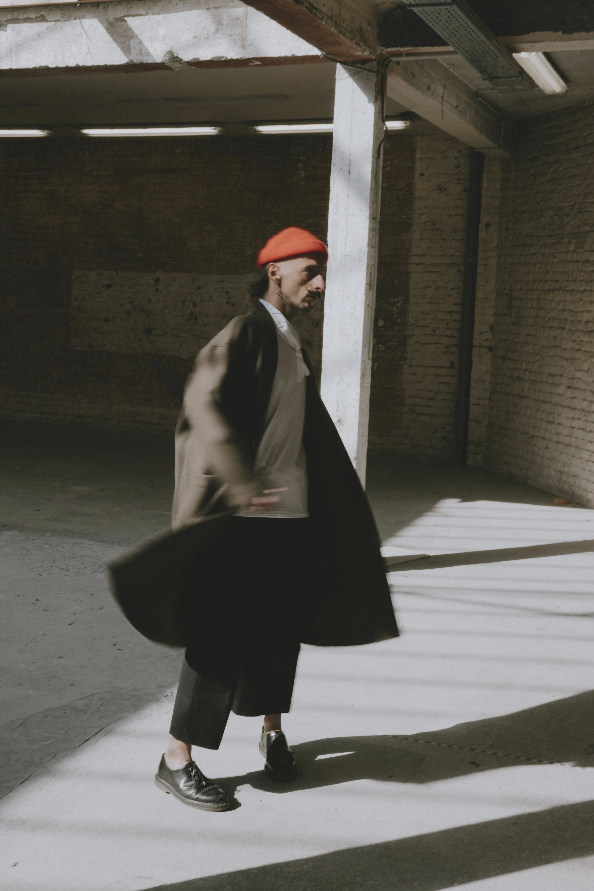 Personne marchant dans une allée urbaine, portant un manteau beige, un pantalon noir, des chaussures noires et un bonnet rouge, avec des ombres portées au sol.