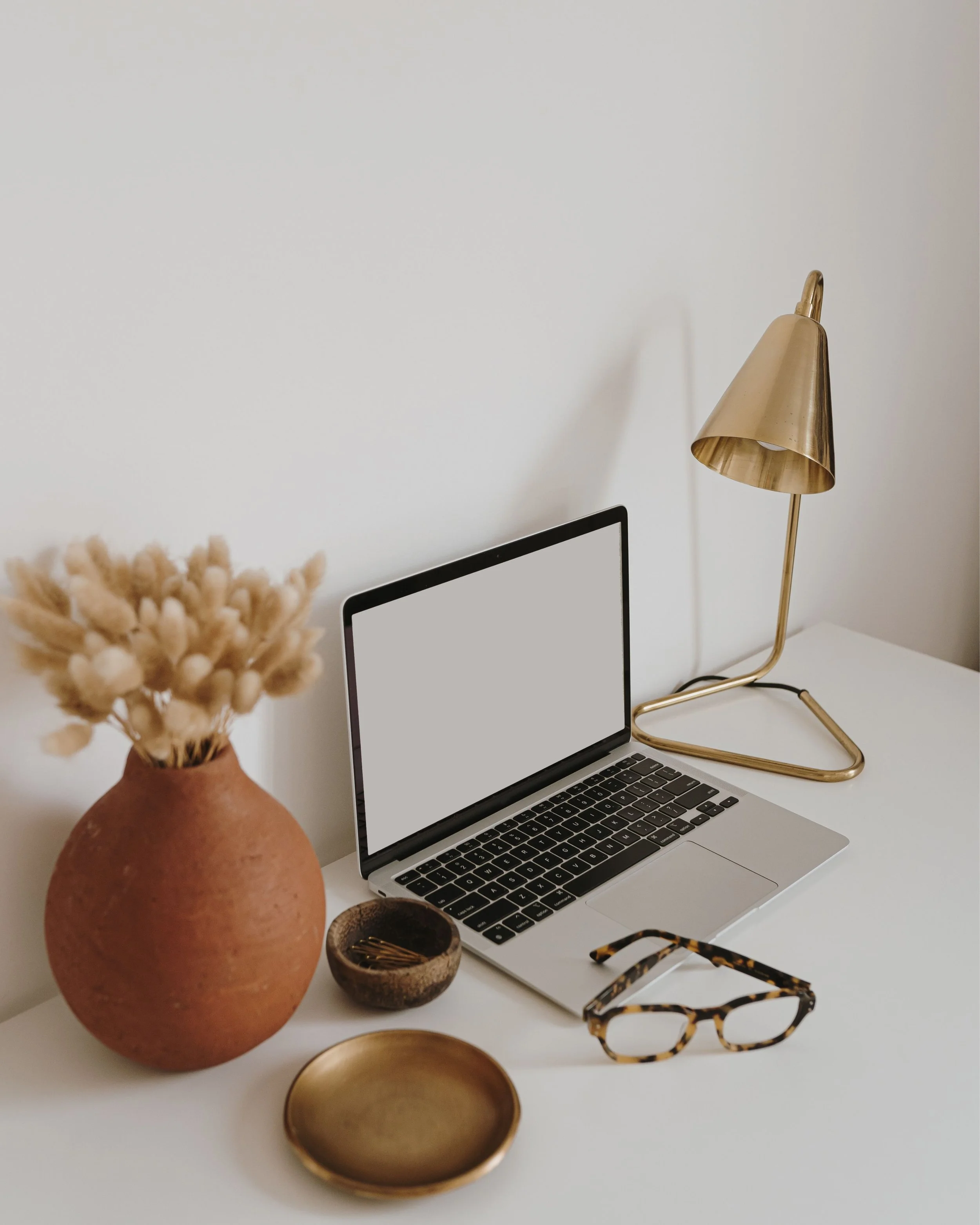 Schreibtisch mit Lampe, Brille, Vase und kleiner Schale am aufgeklappten Laptop