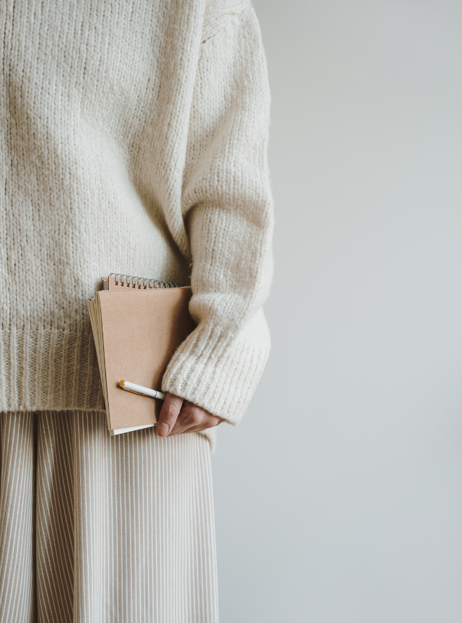 Person trägt einen cremefarbenen Pullover und helle, gestreifte Hosen, hält ein Notizbuch und einen Stift in der Hand.