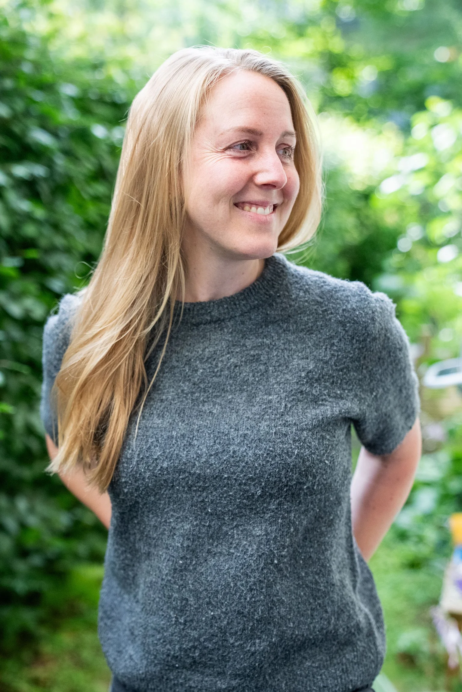 Anna Roth Eine lachende Frau mit langen, blonden Haaren, trägt ein graues T-Shirt und steht im grünen Außenbereich, umgeben von Büschen und Bäumen.