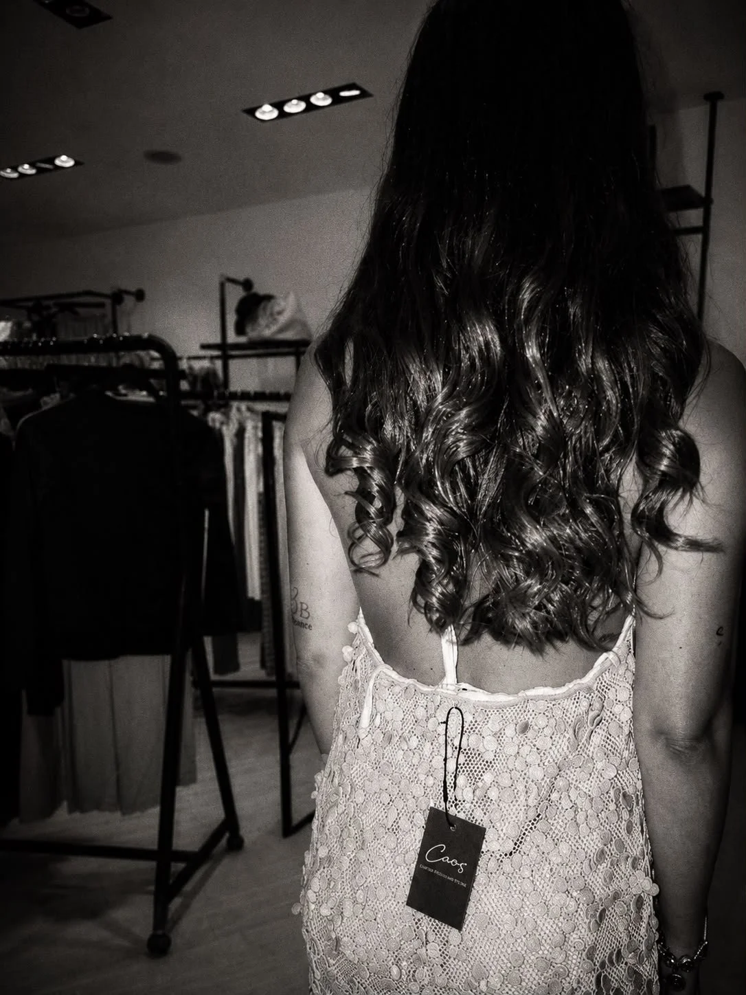 Donna con capelli ricci lunghi in un negozio di abbigliamento. Indossa un vestito in pizzo beige con un'etichetta che dice "Caos".