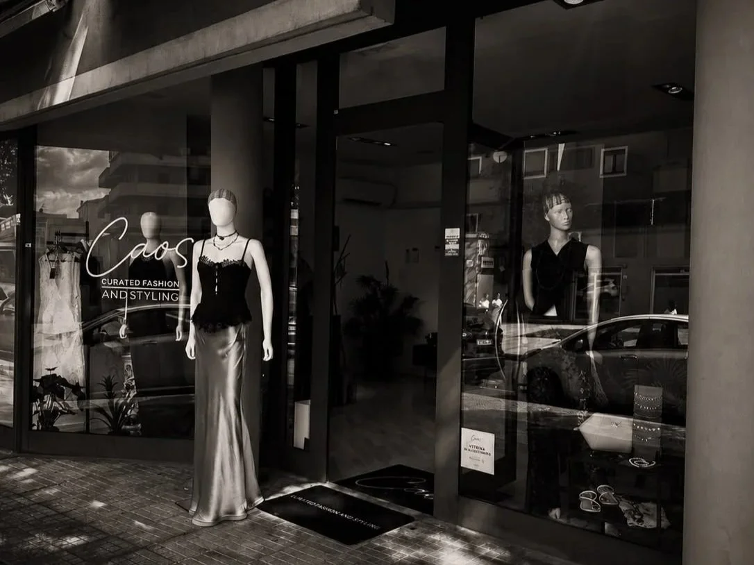Vetrina di un negozio di moda con manichini che indossano abiti eleganti, firma 'Caos' e scritte 'Curated Fashion and Styling'.