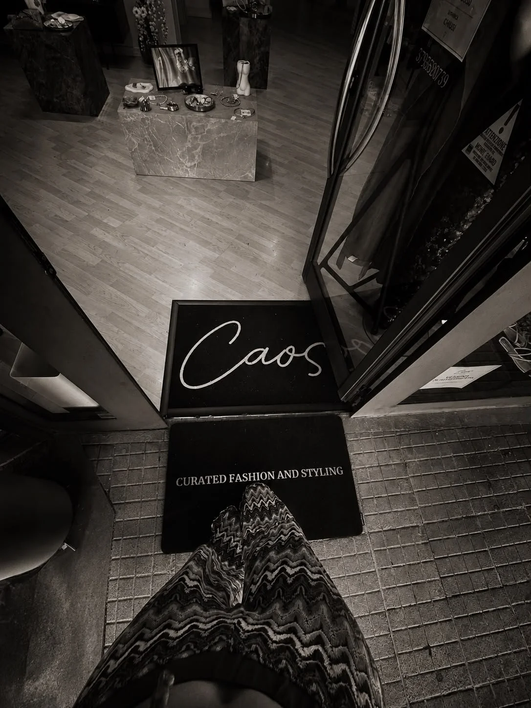 Vista dall'alto di un ingresso di negozio con tappetino nero con scritto "Caos" e altri con scritto "Curated fashion and styling", portone aperto, pavimento in legno e display con gioielli e decorazioni.