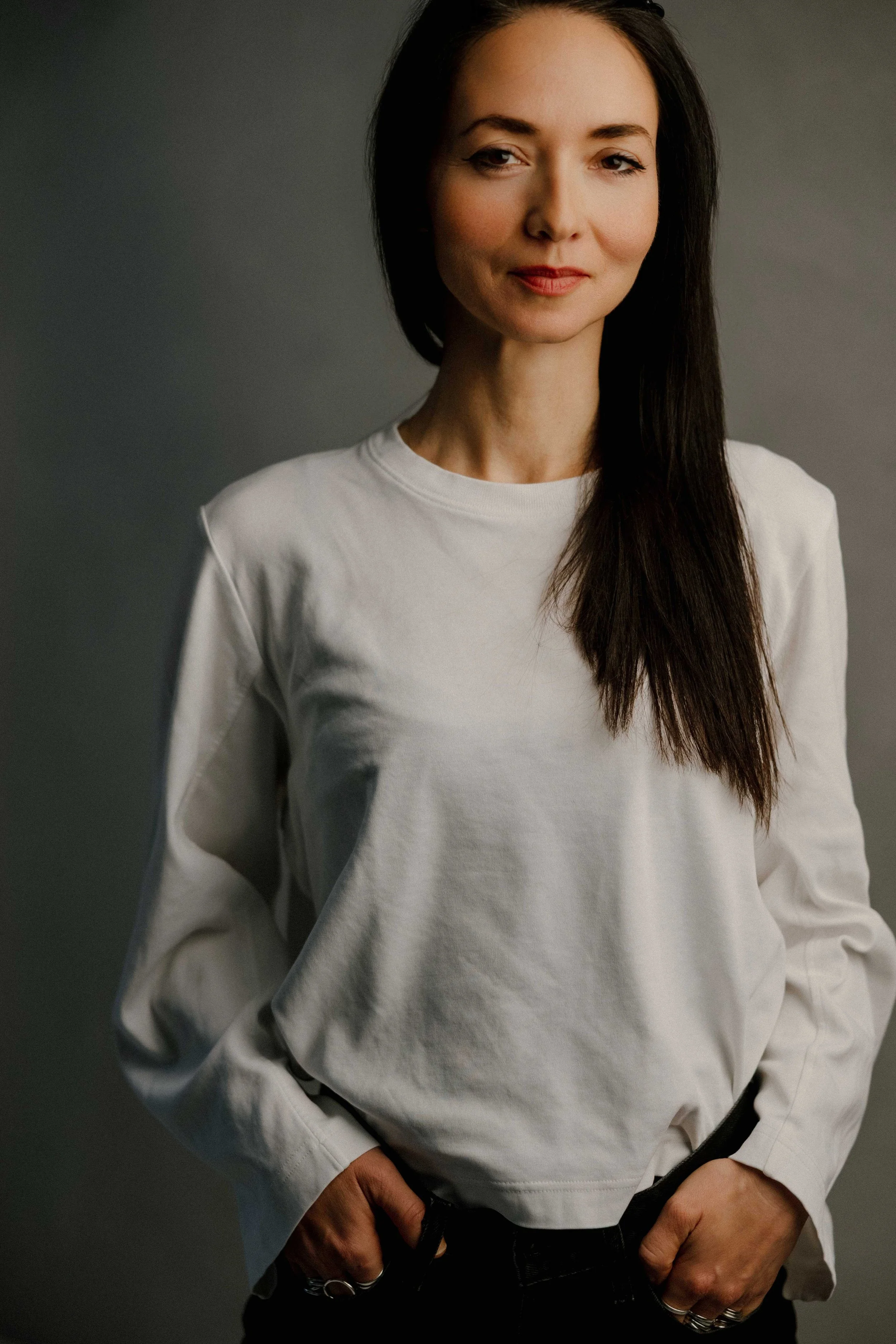 A woman with long dark hair wearing a white long-sleeve shirt, standing against a plain, dark gray background.