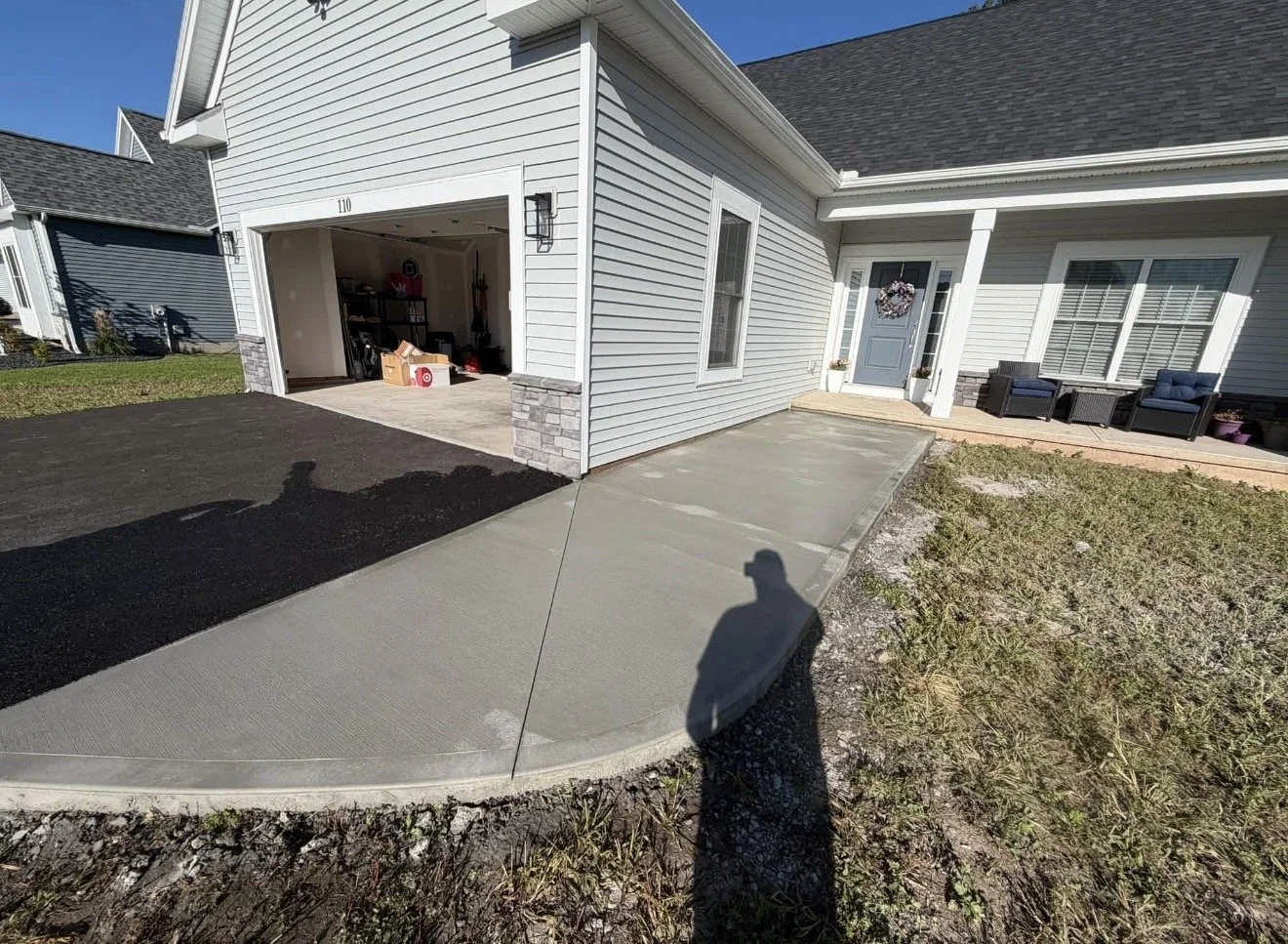Newly poured concrete driveway and sidewalk in front of a house with gray siding and a blue front door, with a garage open showing storage inside. Slightly raised porch with outdoor seating, and a lawn area with sparse grass.